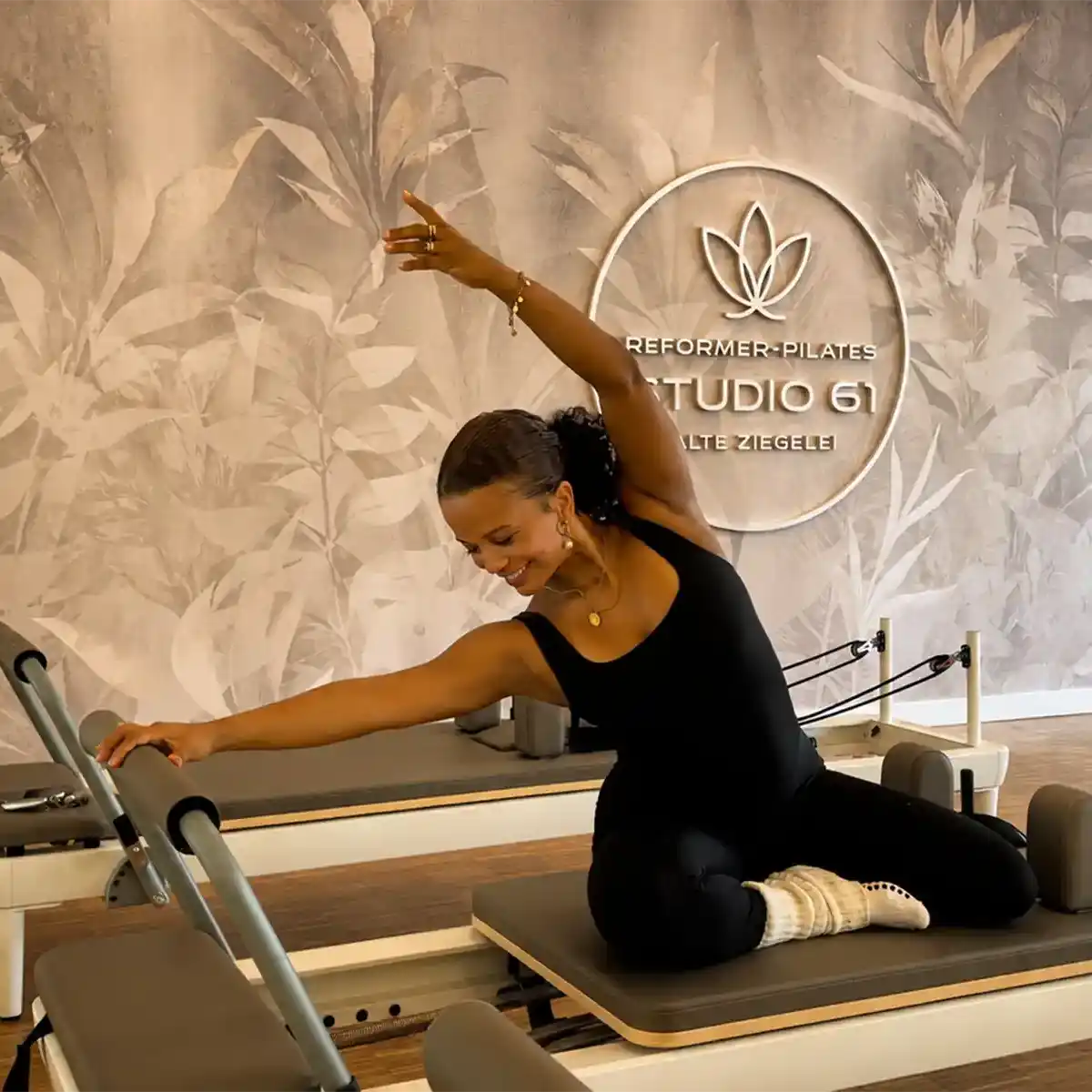 Trainerin in Seitdehnung auf dem Reformer vor dem STUDIO-61-Logo – Training für Haltung und Körpergefühl